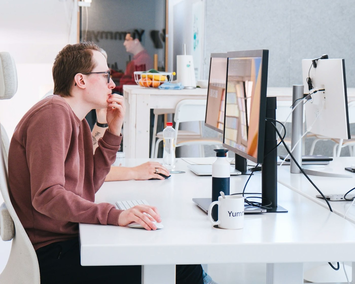 White office with person in red brown sweater in focus, as seen from the side. The person is looking at their computer monitor. In the rear is a person, out of focus, behind a glass door.