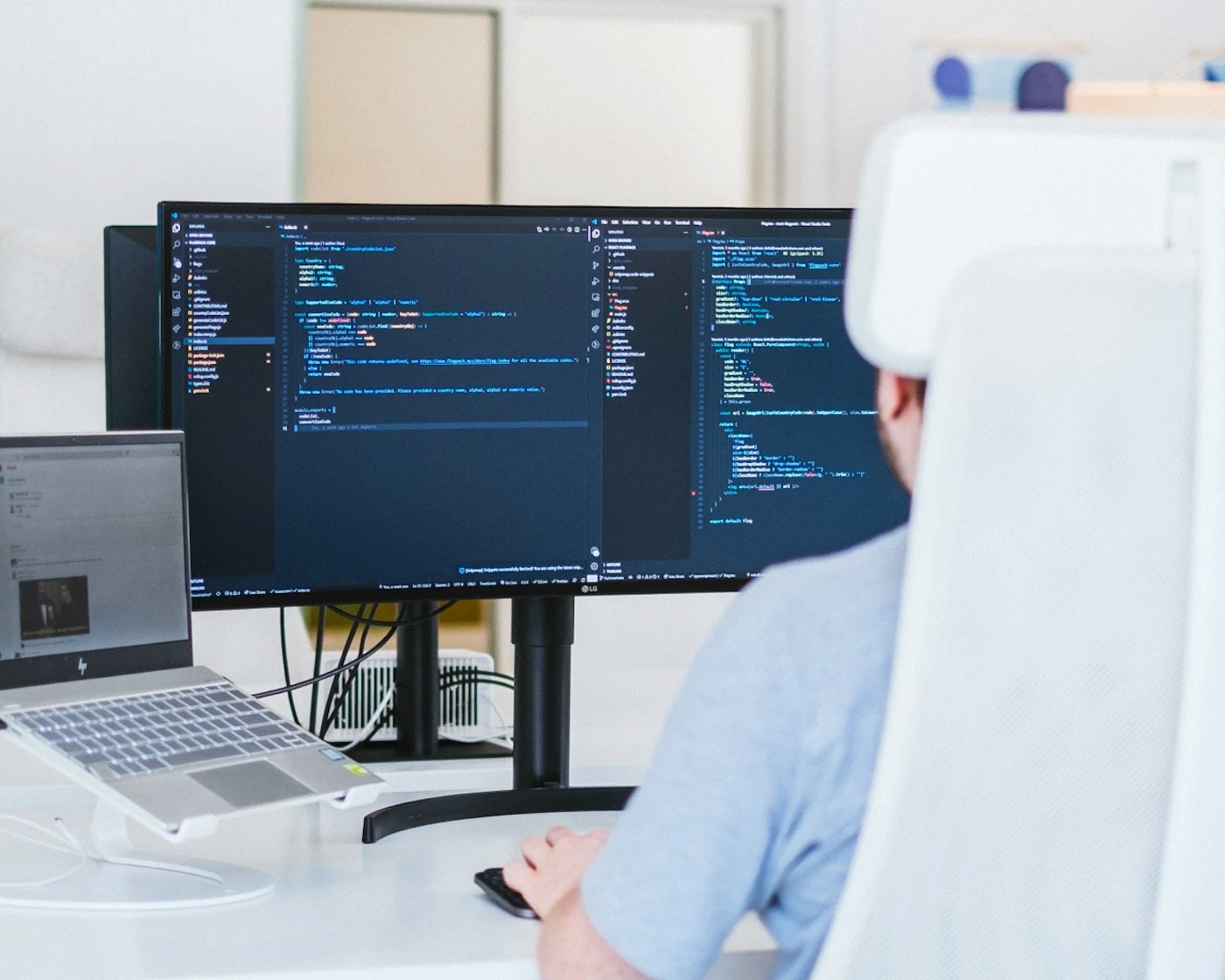 Office chair with a person in it as seen from behind. The person is sitting behind a computer monitor that shows a dark blue code editor. On the left of the computer monitor is an laptop on a laptop stand.