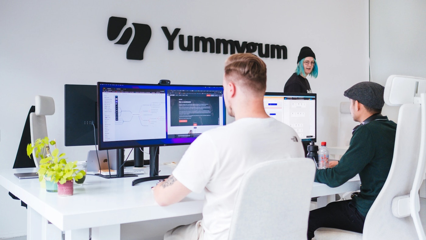 People sitting behind their computer monitors, in a white office. In the rear is a large blak logo that reads Yummygum.