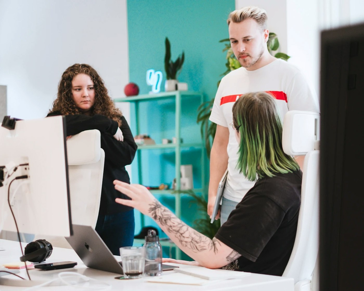 One person sitting beind a computer, hand gesturing as they're speaking to two other people who are standing and looking at the first mentioned person. In the background is a turquoise background.