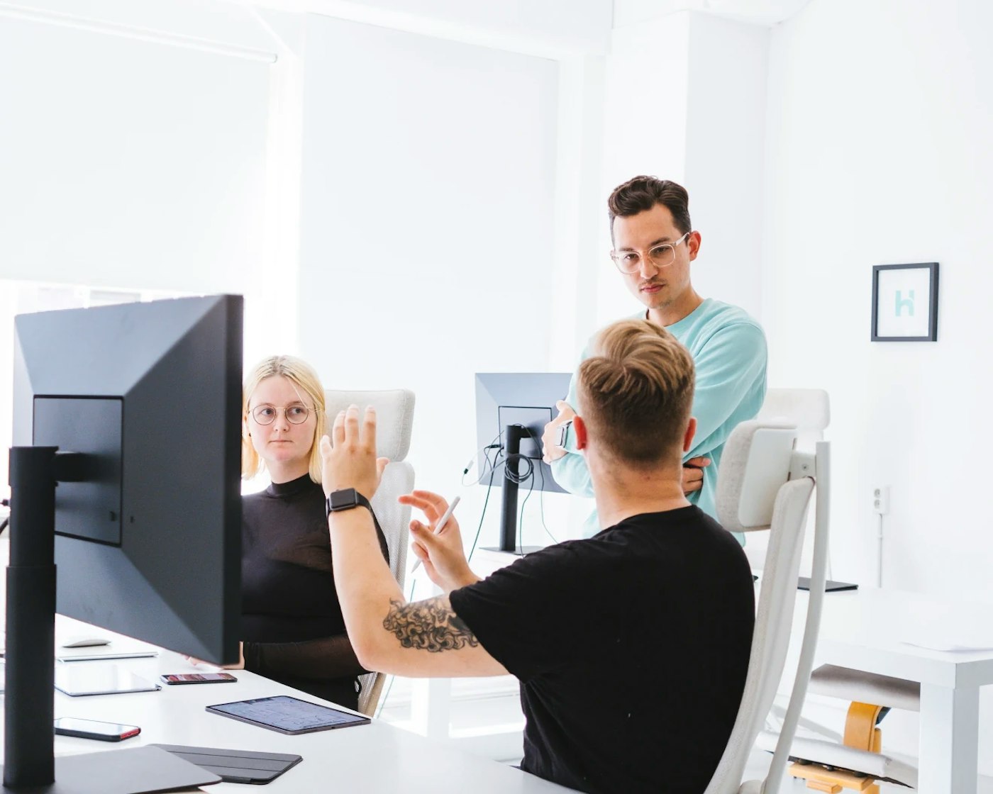 Three people in a white interior office having a conversation. Two of them are sitting behind a desk while the third one is standing.