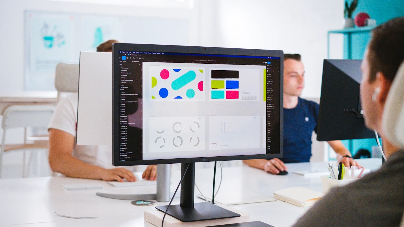 Person looking at computer monitor that shows bright colorful shapes. On opposing side of the first person are two persons working behind their respective computer.