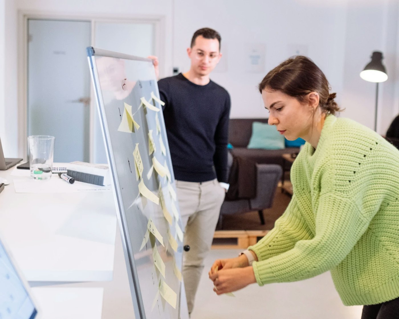 Person reaching towards a whiteboard that shows post-its to add a yellow post-it to the white board. In the rear is a person observing the action.