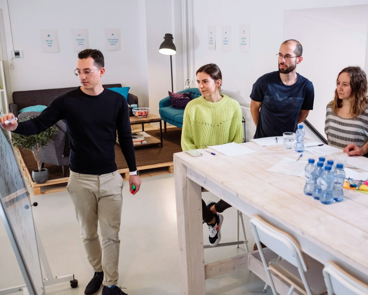 Group of people all looking at a white board that is directed towards them. One person is standing besides the whiteboard while drawing something on the whiteboard using a marker. The other people are gathered around a high top table.