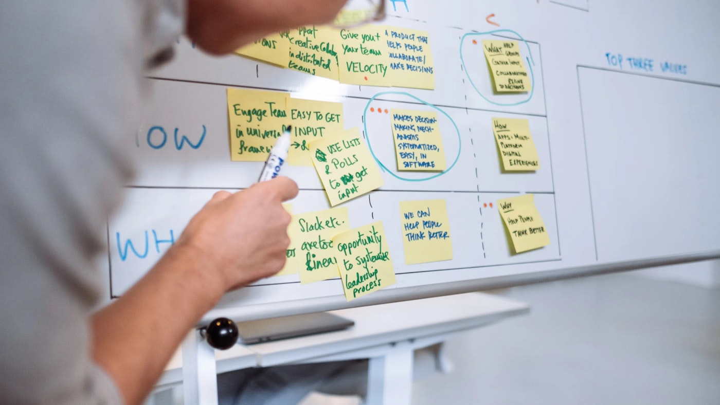 Person hovering in front of white board, while point a marker at yellow sticky notes that have text on them, and are placed on a grid.