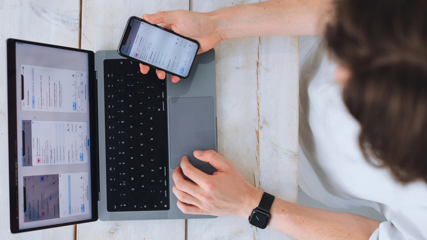 Person with one hand resting on laptop and the other hand holding a smart phone that shows a user interface, as photographed from above.