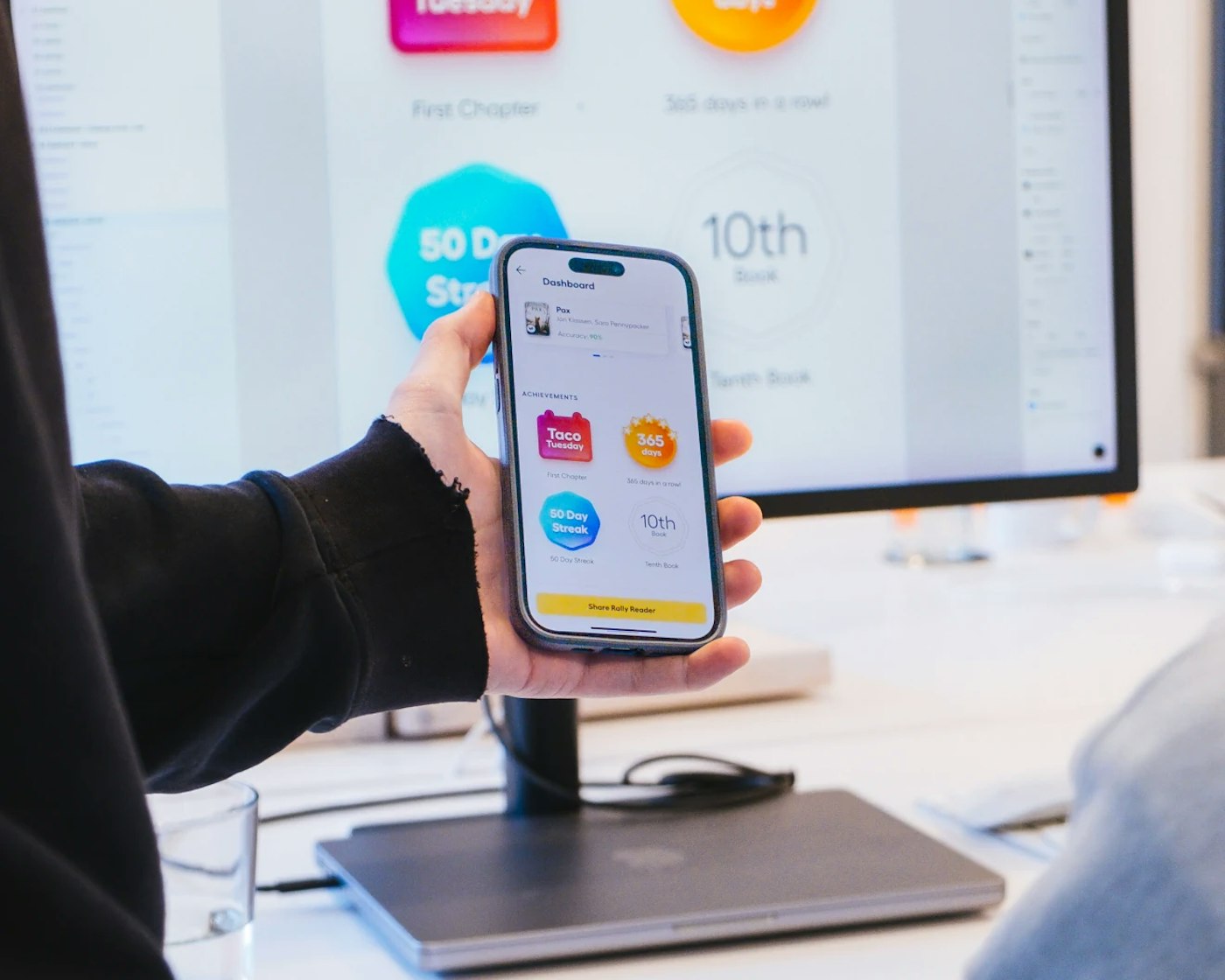 Hand holding a smart phone that shows a user interface with a white background and colorful shapes. Behind their hand is a large computer monitor that shows a similar user interface zoomed in.