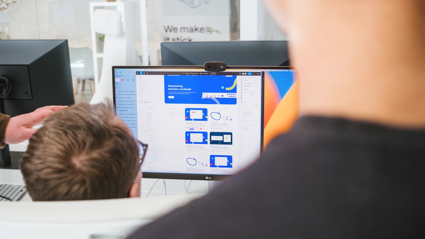 Person as seen from behind, looking at their computer monitor. The computer monitor shows a white window with blue cards. On the right is someone's neck and shoulder, out of focus.