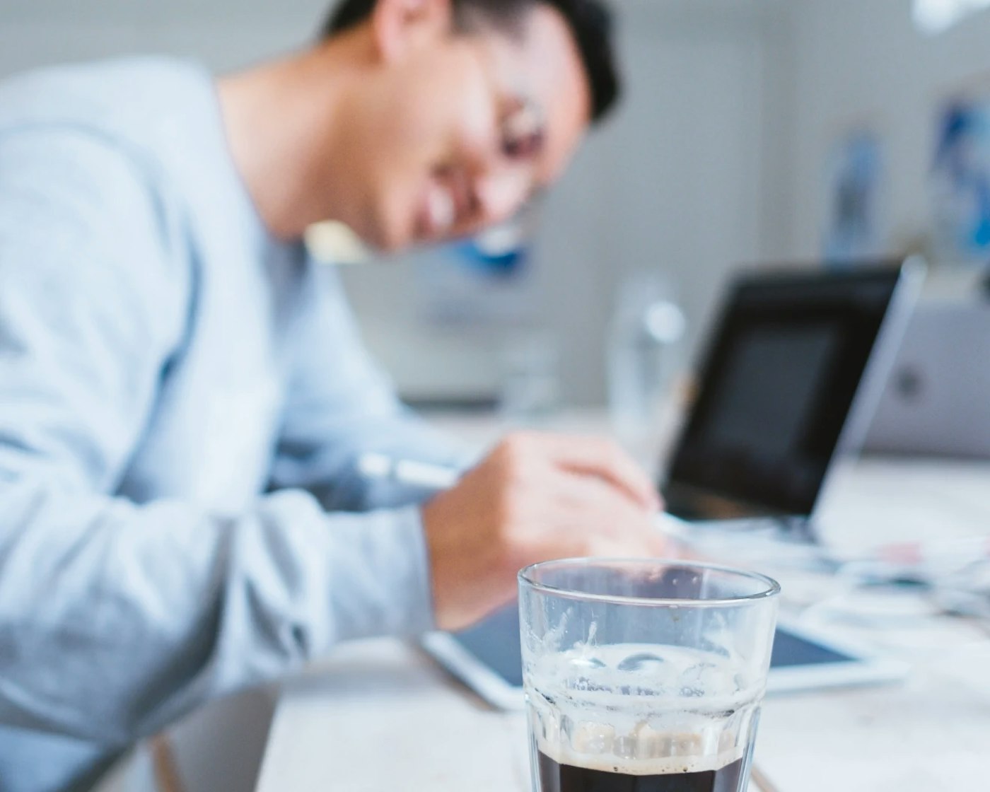 Person out of focus, smiling while controling their ipad. Close to the camera, in focus, is a black cup of coffee.