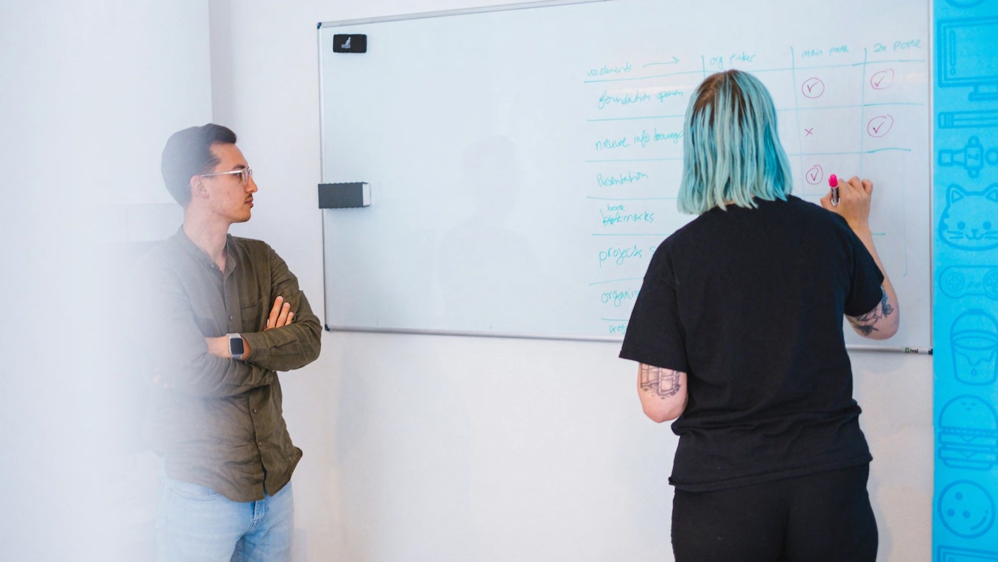 One person with arms crossed looking at another person who is writing something on a whiteboard using a marker.