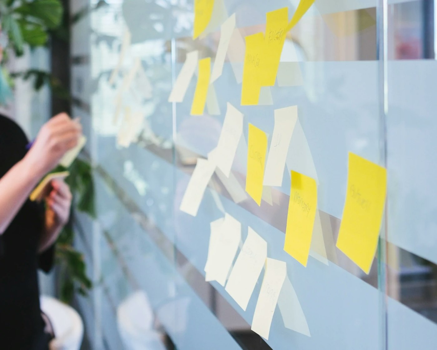 Glass wall seen from the side, with a dozen yellow post its on it. In the rear is a person's arm showing just within the crop, sticking more post its on the glass wall.