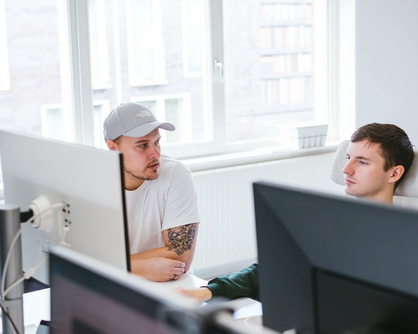 Two people sitting side by side behind a set of computer monitors. One of them is looking at the other.