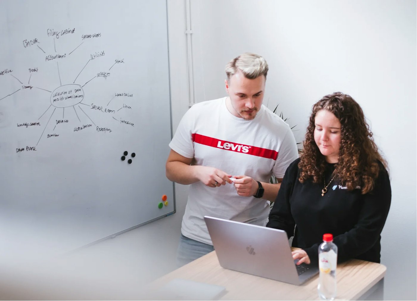 Two persons standing next to a whiteboard that has mindmap drawn on it with a marker. Both persons standing side by side while both of them are looking one laptop.