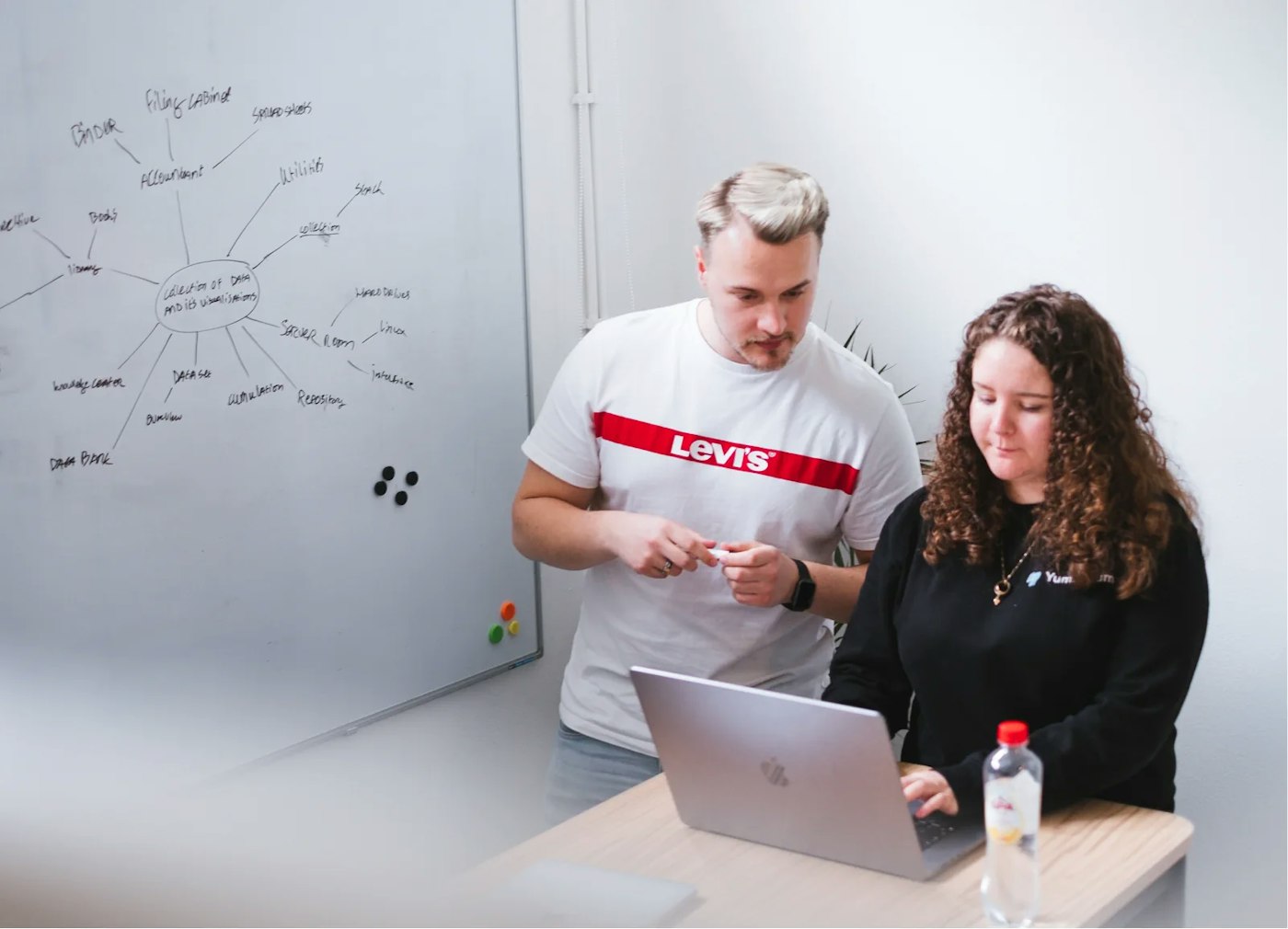 Two persons standing next to a whiteboard that has mindmap drawn on it with a marker. Both persons standing side by side while both of them are looking one laptop.