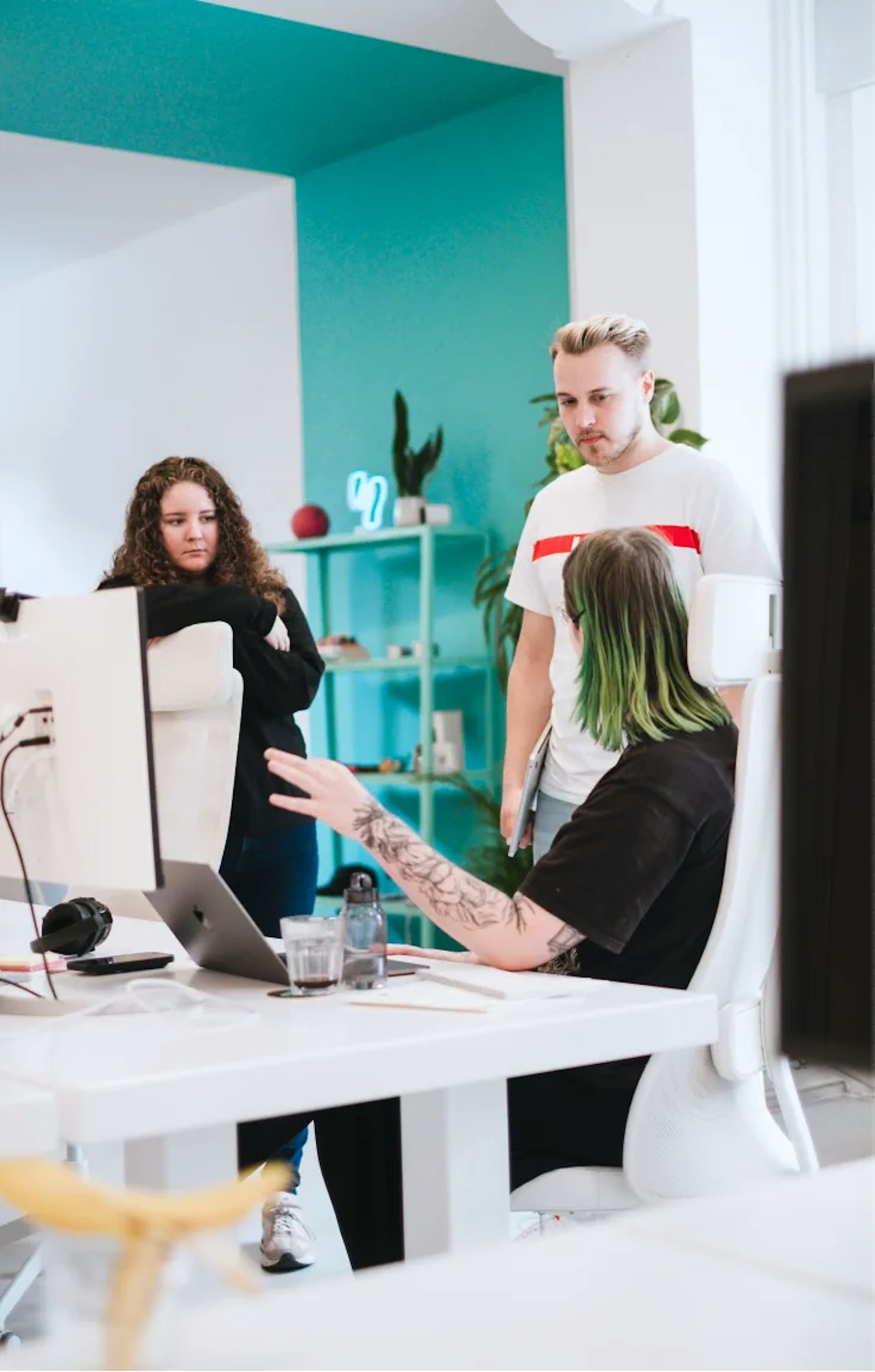 Three people of which two are standing and looking at the third person who is sitting. Person who is sitting is gesturing with their hands, as if their explaining something to the two standing persons. In the background is a partially turquoise colored wall and ceiling.