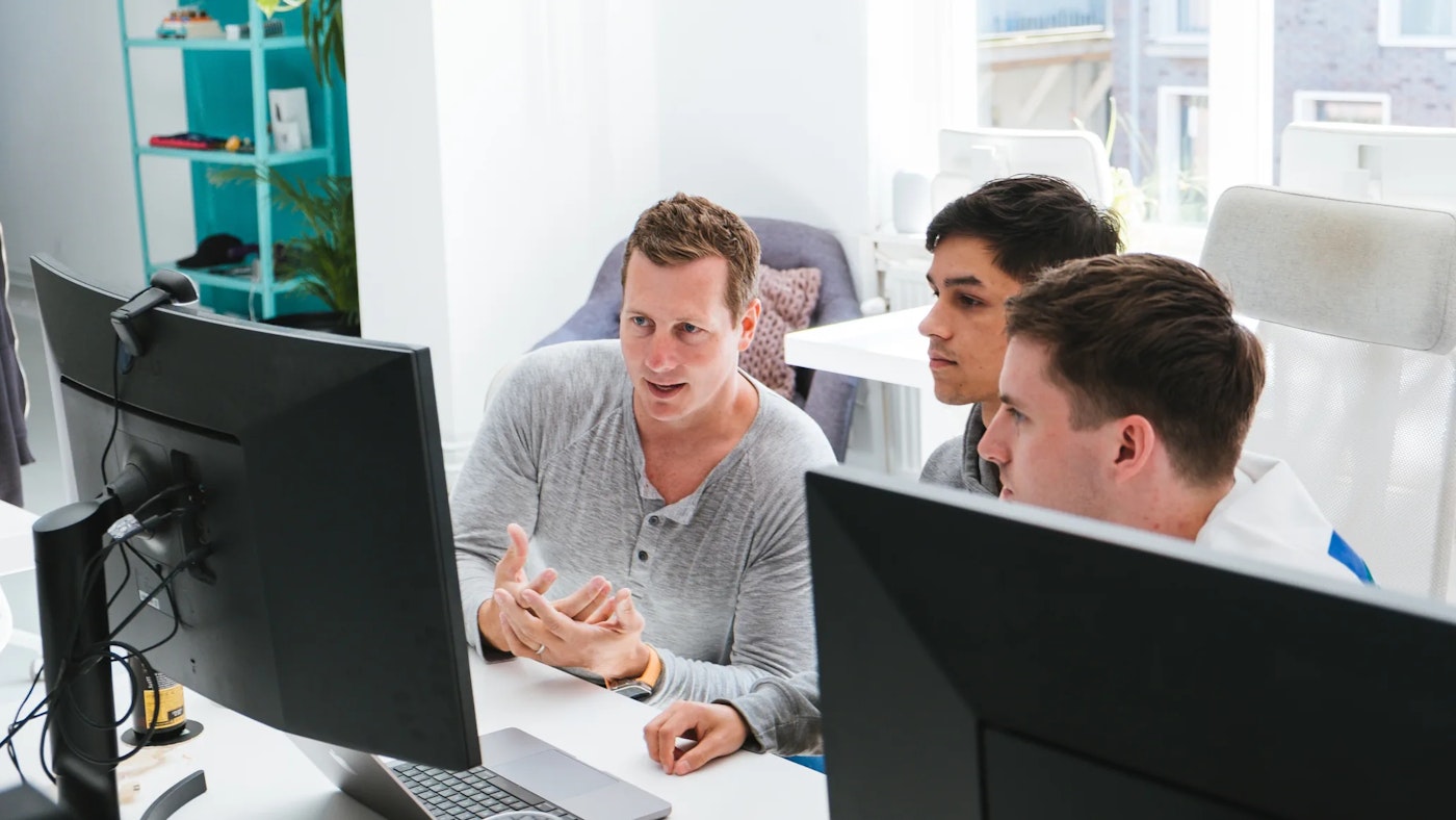 Three people sitting behind computer monitors. Two of them are looking at each other while one is gesturing with their hands, seemingly explaining something to the other..