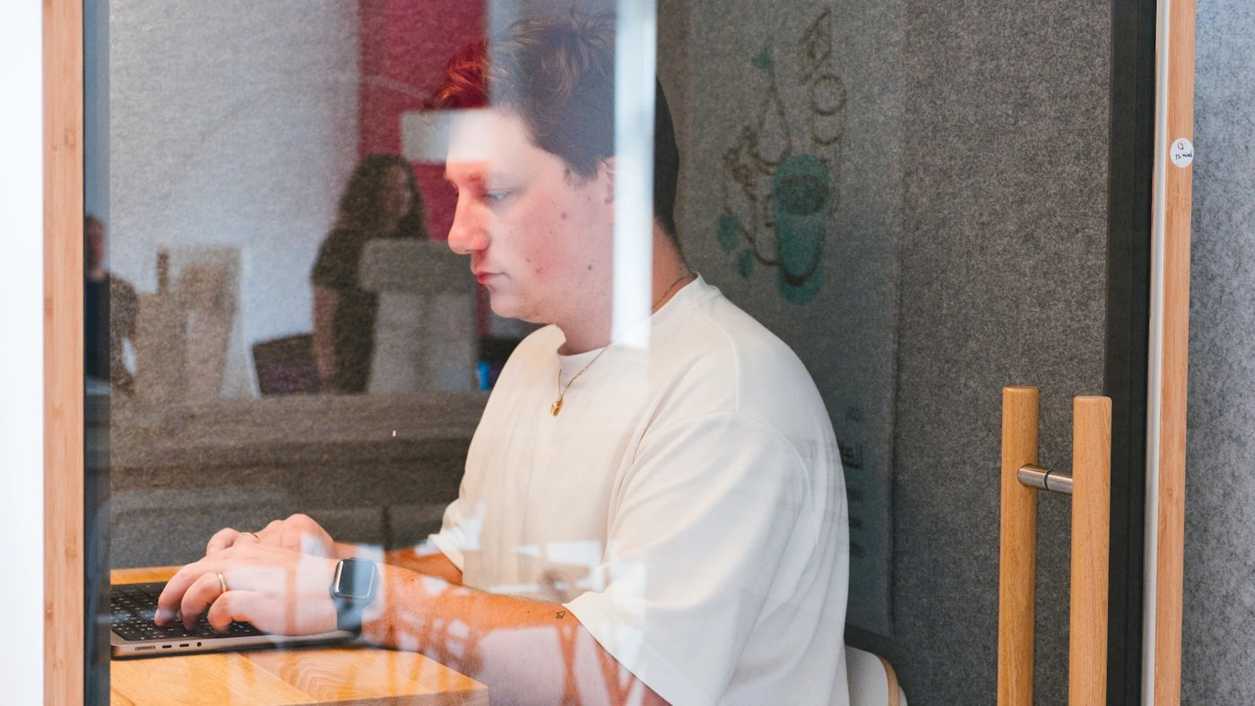 Person as seen through glass door of an acoustic booth, working on their laptop that is placed on a wooden shelf. Interior of the booth is dark gray. The reflection of the glass door shows a person walking.