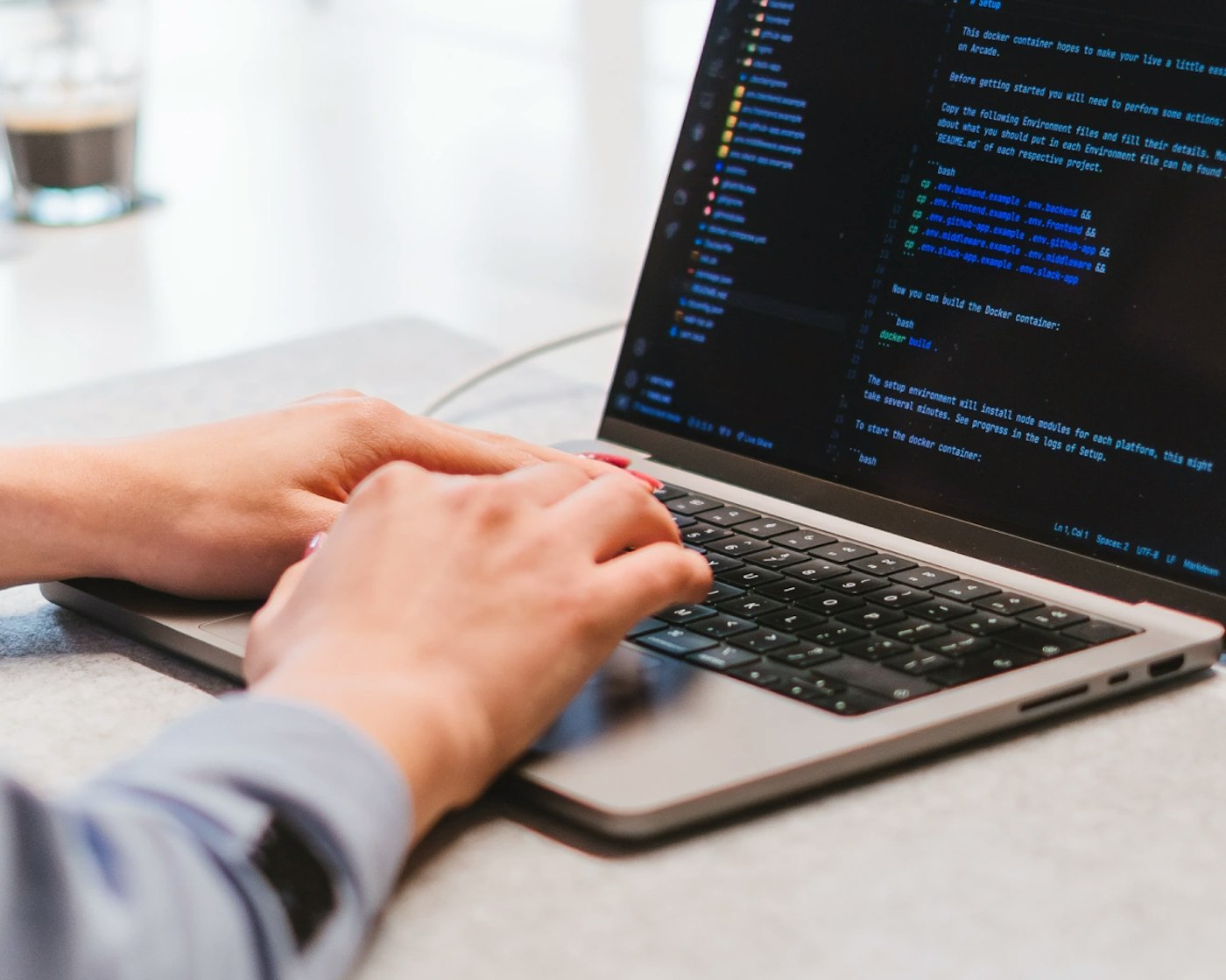 Close up of two hands typing on a laptop keyboard. The laptop's display shows a dark themed code editor.