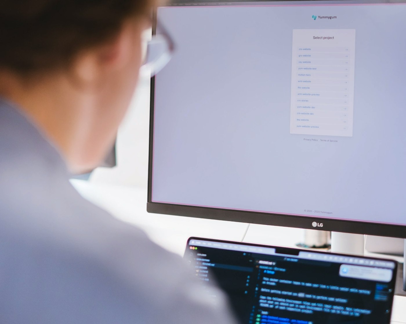 Person sitting behind a display and a laptop. The display shows a list of items whereas the laptop shows a code editor.