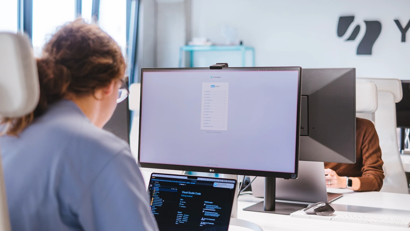 Person sitting behind a display and a laptop in an office space. The display shows a list of items whereas the laptop shows a code editor.