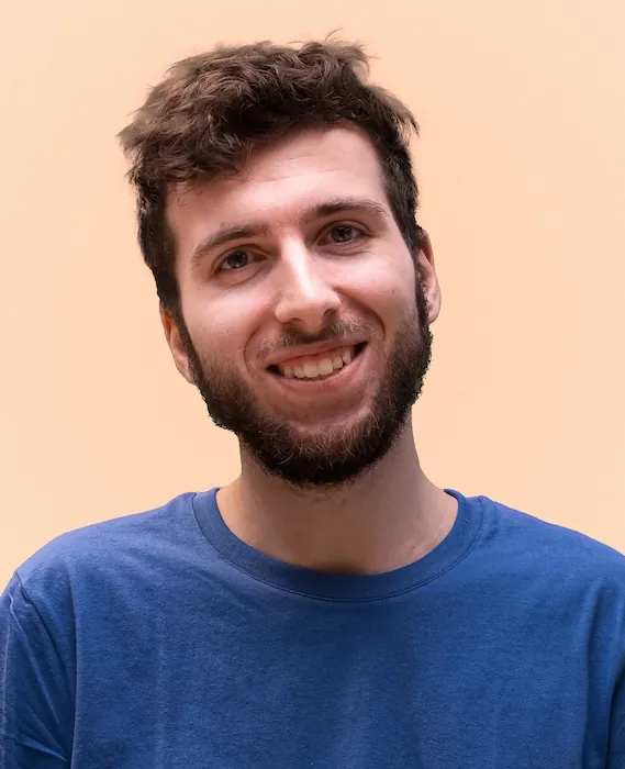 Portrait of person dressed in a blue t-shirt, against a peach colored backdrop.