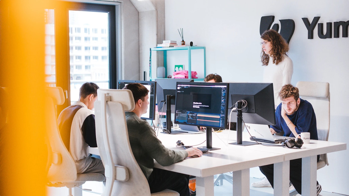 Group of five people in a mainly white interior office. Four of them are sitting at their desk, working on a computer screen while one of them is standing. On the left out of focus there's a bright yellow blurred line.