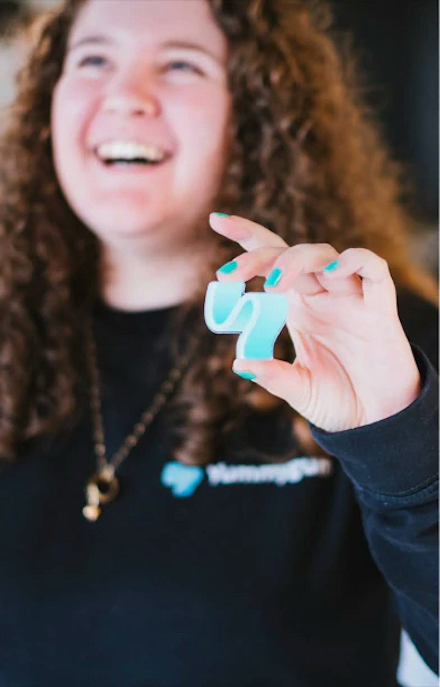 Smiling person holding up an enamel pin of a turquoise Yummygum logo mark.
