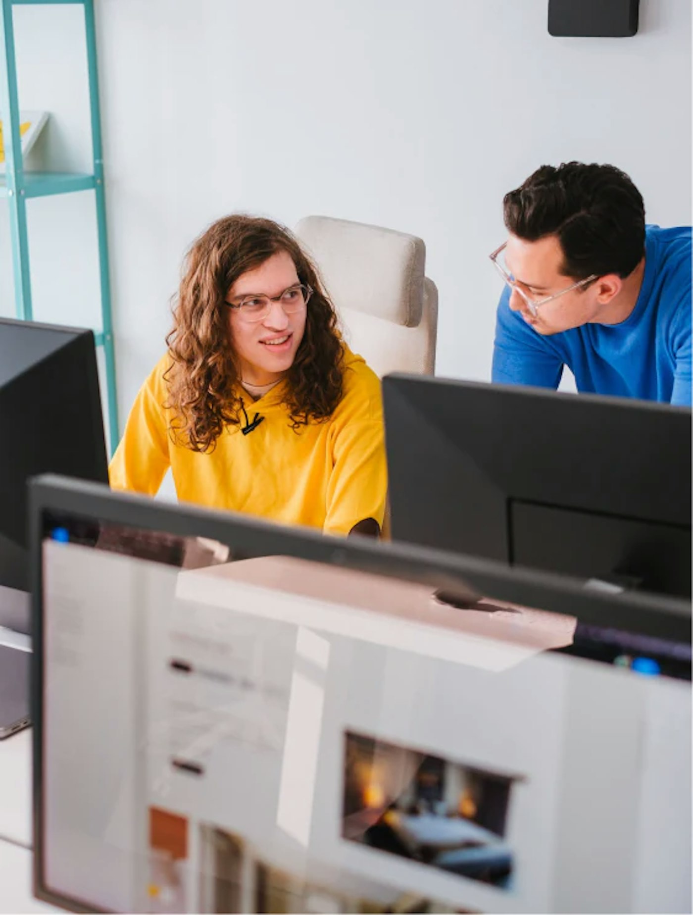 Two people having a conversation behind a desk, as seen behind a set of computer displays. One of them is wearing a bright yellow sweater and the other one is wearing a bright blue sweater.