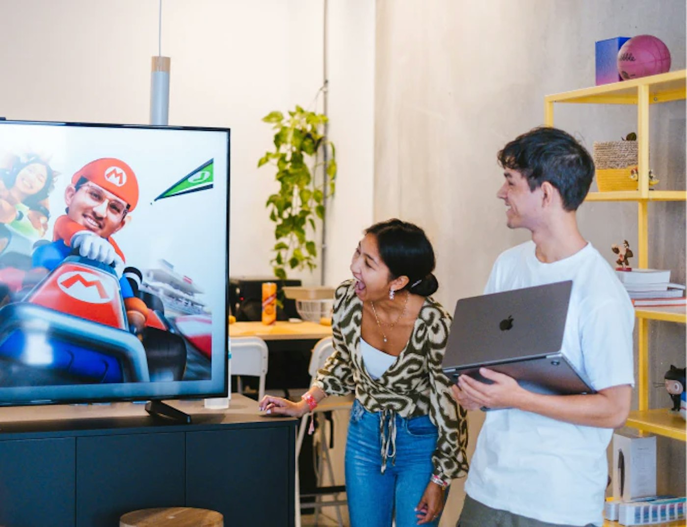 Two people looking and laughing at a television screen that shows a Mario Kart video game character with a face of a Yummygum team member pasted onto it.
