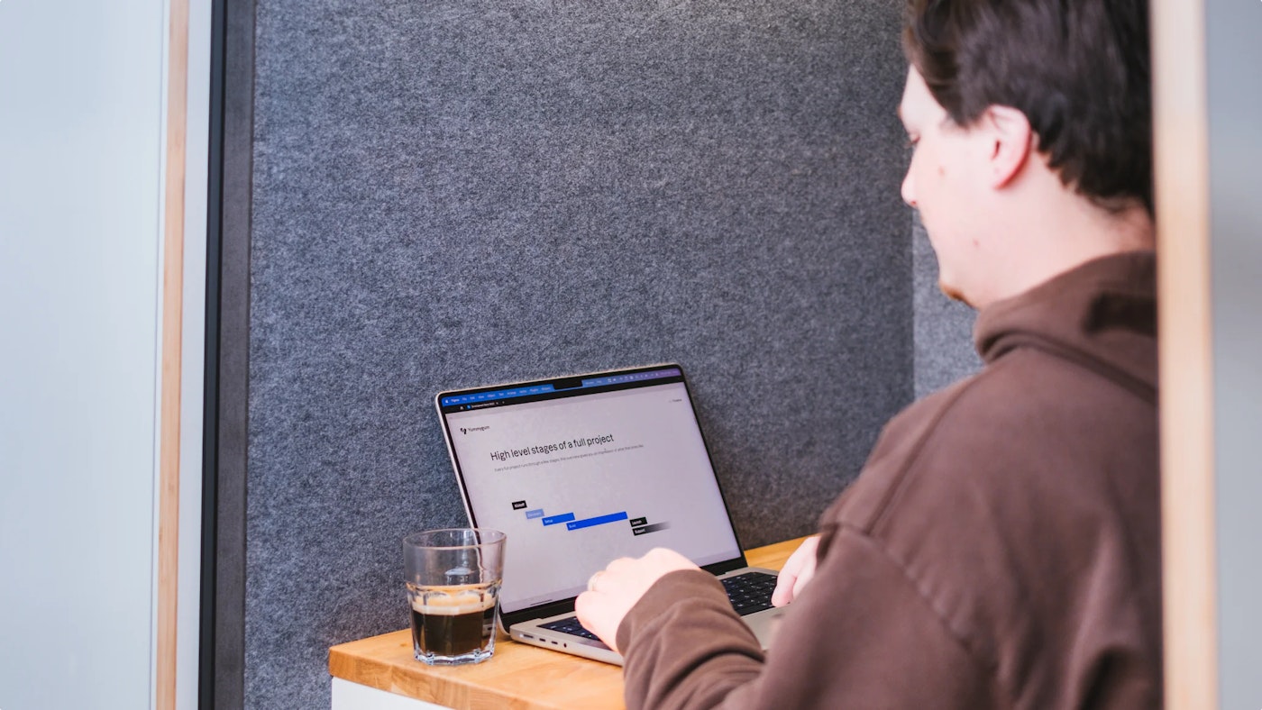A person (Head of Operations Donovan, writer of this blog post) in a brown sweatshirt working on a laptop in a small booth. The laptop screen shows a project timeline labeled "High level stages of a full project" with phases marked as "Start," "Development," and "Finish." A glass of coffee is placed on the table beside the laptop.