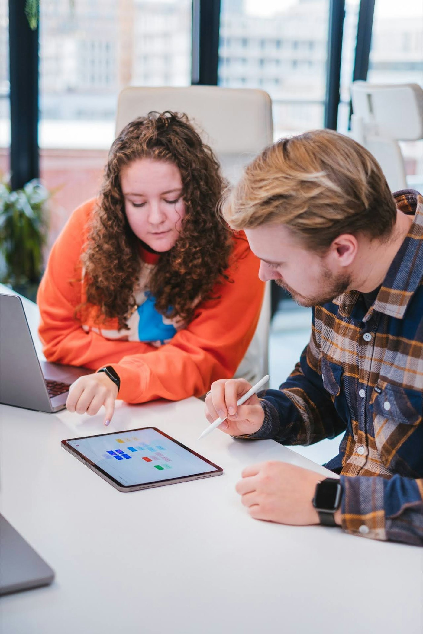 Two people collaborating at a table with laptops. One is using a stylus on a tablet displaying colorful charts. They are in a modern office setting with large windows in the background.