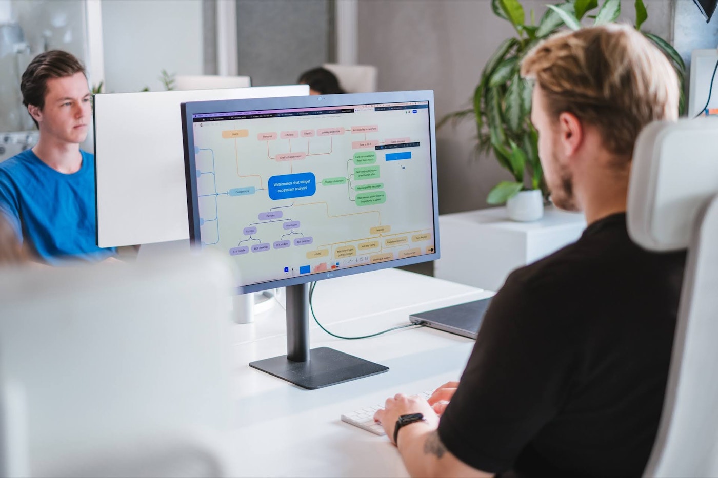 A man sits at a desk working on a computer displaying a colorful flowchart. Another person in a blue shirt sits at a separate desk. The workspace is modern with white furniture and plants in the background.