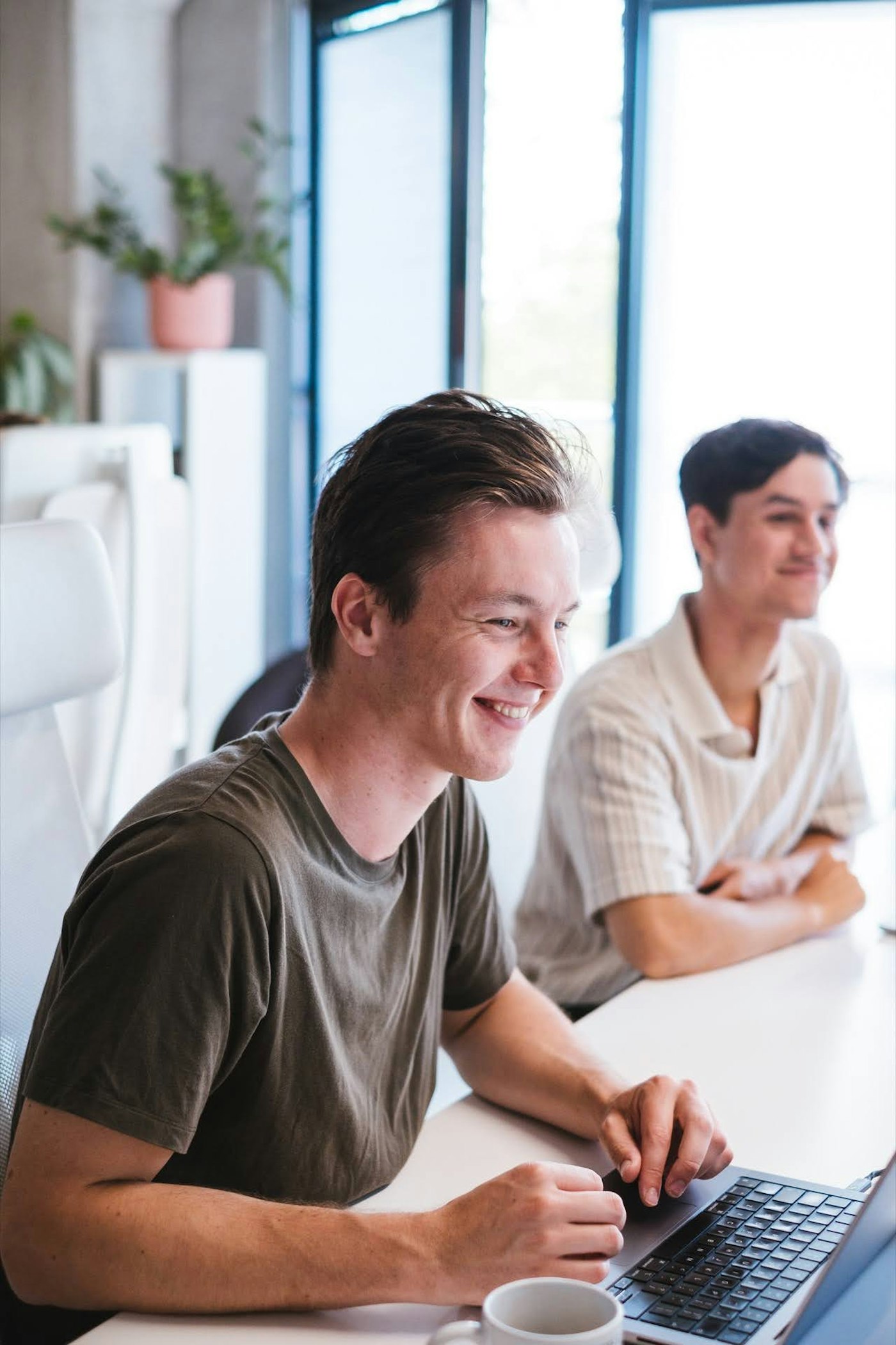 Two young men are sitting at a table in a bright room with open laptops. They are smiling and appear engaged. One wears a dark t-shirt and the other a striped shirt. A plant and large windows are visible in the background.