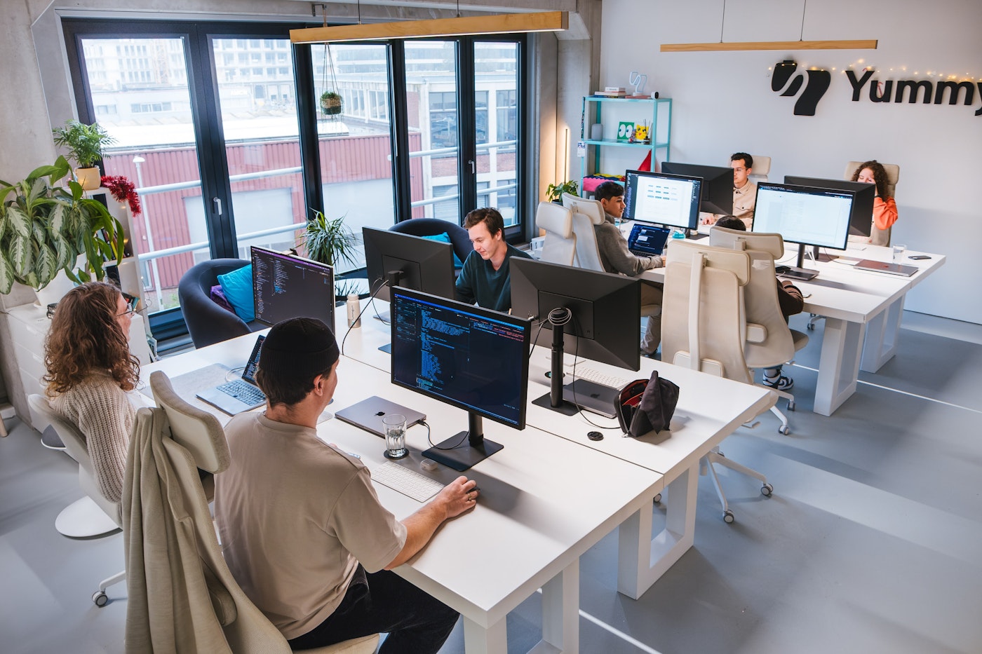 A modern office scene with six people working at desks with computers. Large windows let in natural light, and there are plants around. The atmosphere is collaborative and focused.