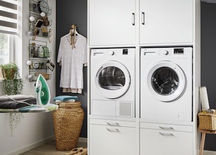 Hierbij een voorbeeld van een veilig opgestelde wasmachine en droger, die verhoogd zijn geplaatst in een witte kast in een waskamer.