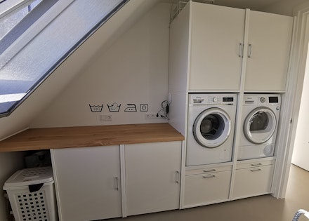 Wasmachine en droger hoger geplaatst voor de ideale werkhoogte, onder een schuin dak met genoeg opbergruimte voor de wasmand, onderkasten, lades en uittrekbare plank