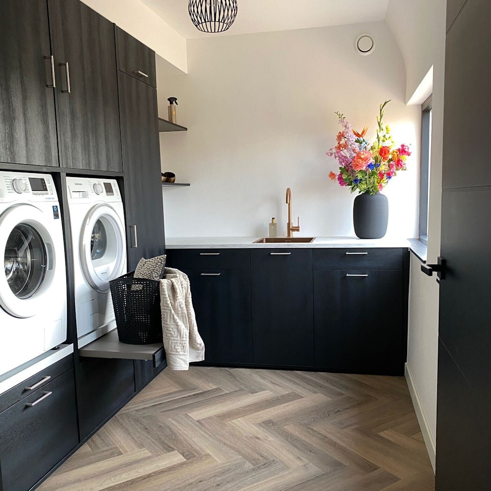 Waschküche in L-Form mit schwarzem Waschmaschinenschrank und Ausziehplatte für Wäschekörbe.