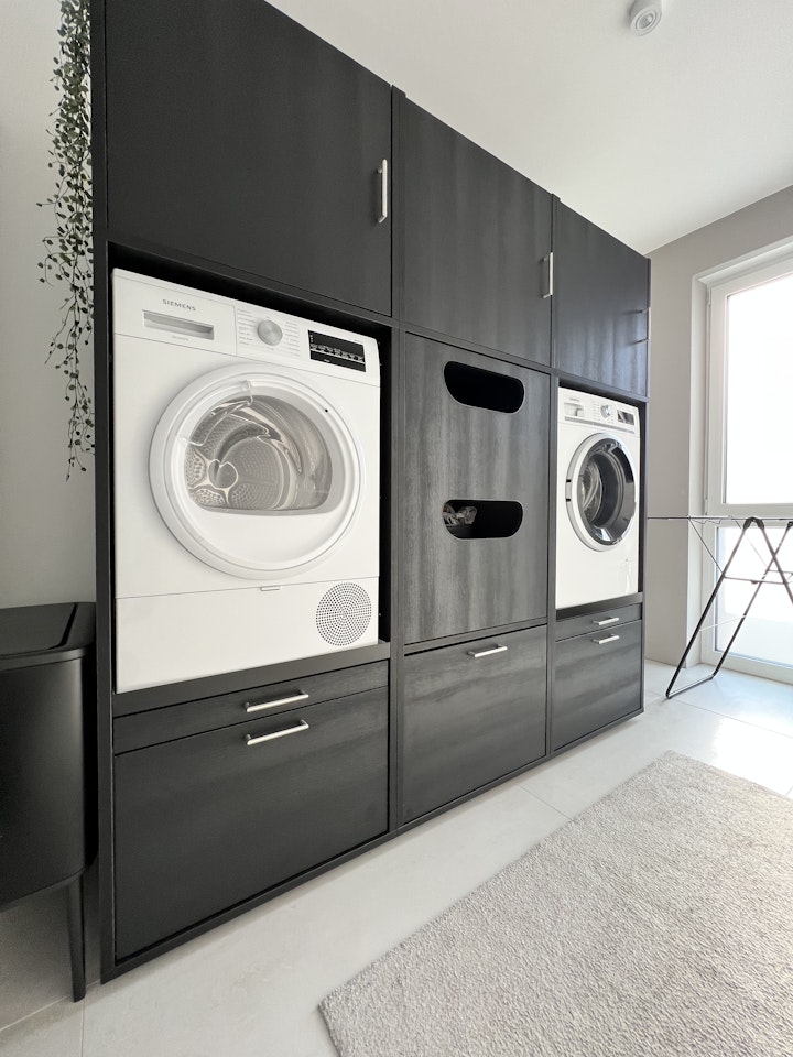 Idee voor berging/washok waarbij wasmachine en droger in een zwarte wasmachine kast zijn geplaatst. Met exta opbergruimte en kast voor de wasmand.