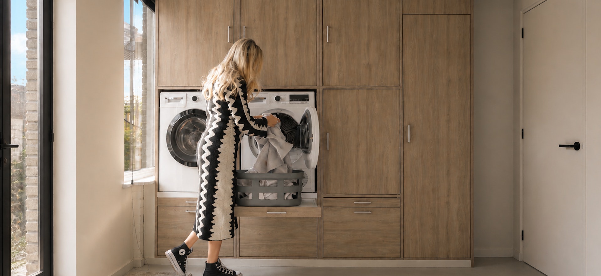 Een vrouw in een zwart-wit gestreepte jurk plaatst schone was in een wasmachine, terwijl een wasmand met kleding op een kastje naast de machine staat. De ruimte is modern ingericht met houten kasten en grote ramen die natuurlijk licht binnenlaten.