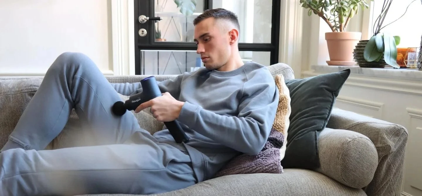 En man som ligger i soffan men gråa mjukiskläder och masserar sitt ben med en flowgun, massagepistol