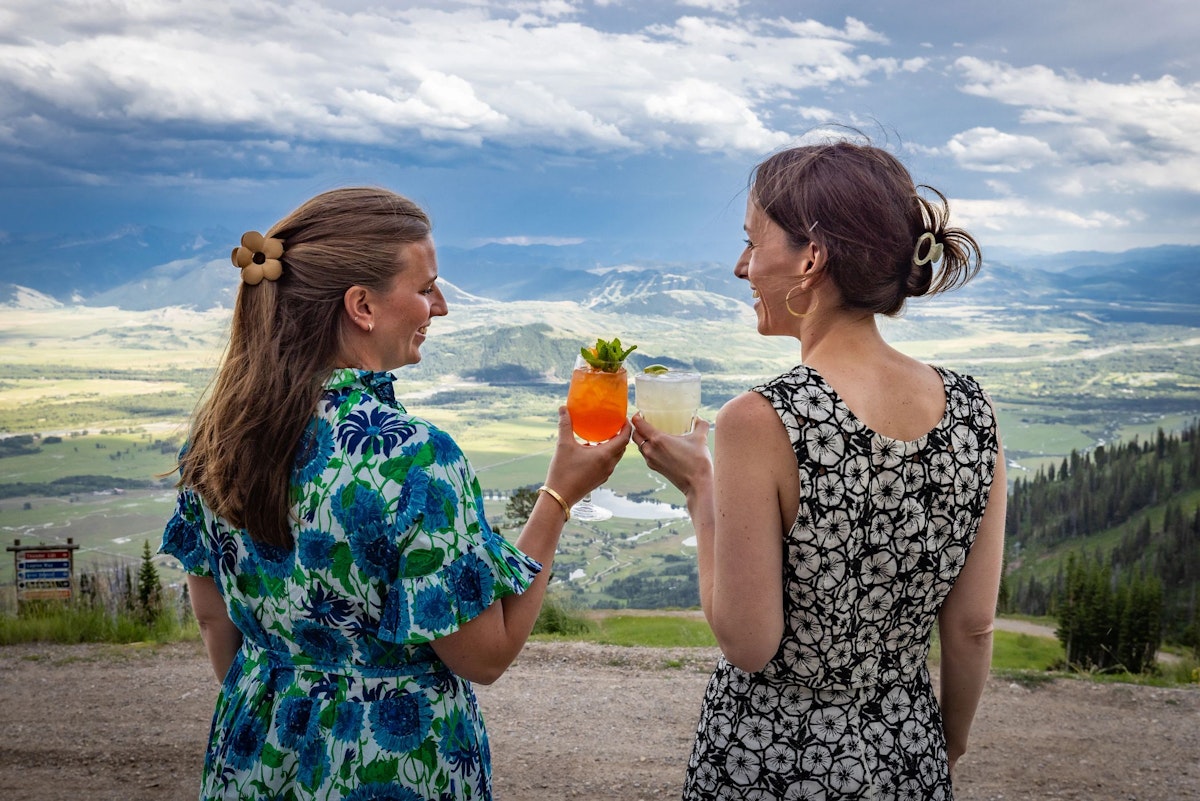 Two women clinking cocktails with the view from the Deck at Piste