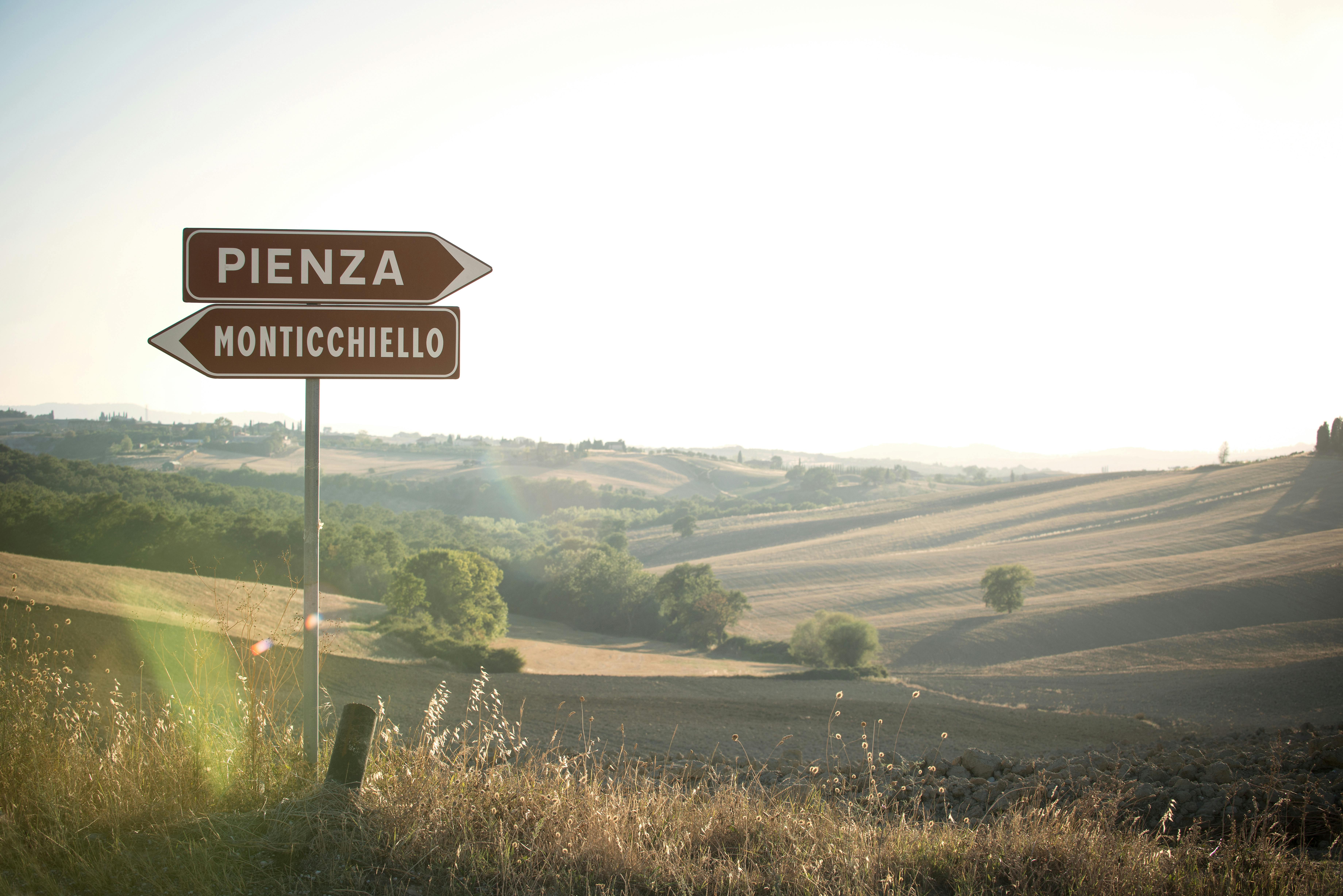 Two brown road signs of Pienza toward the right and Montichiello towards the left with Val d'Orcia landscape at sunset
