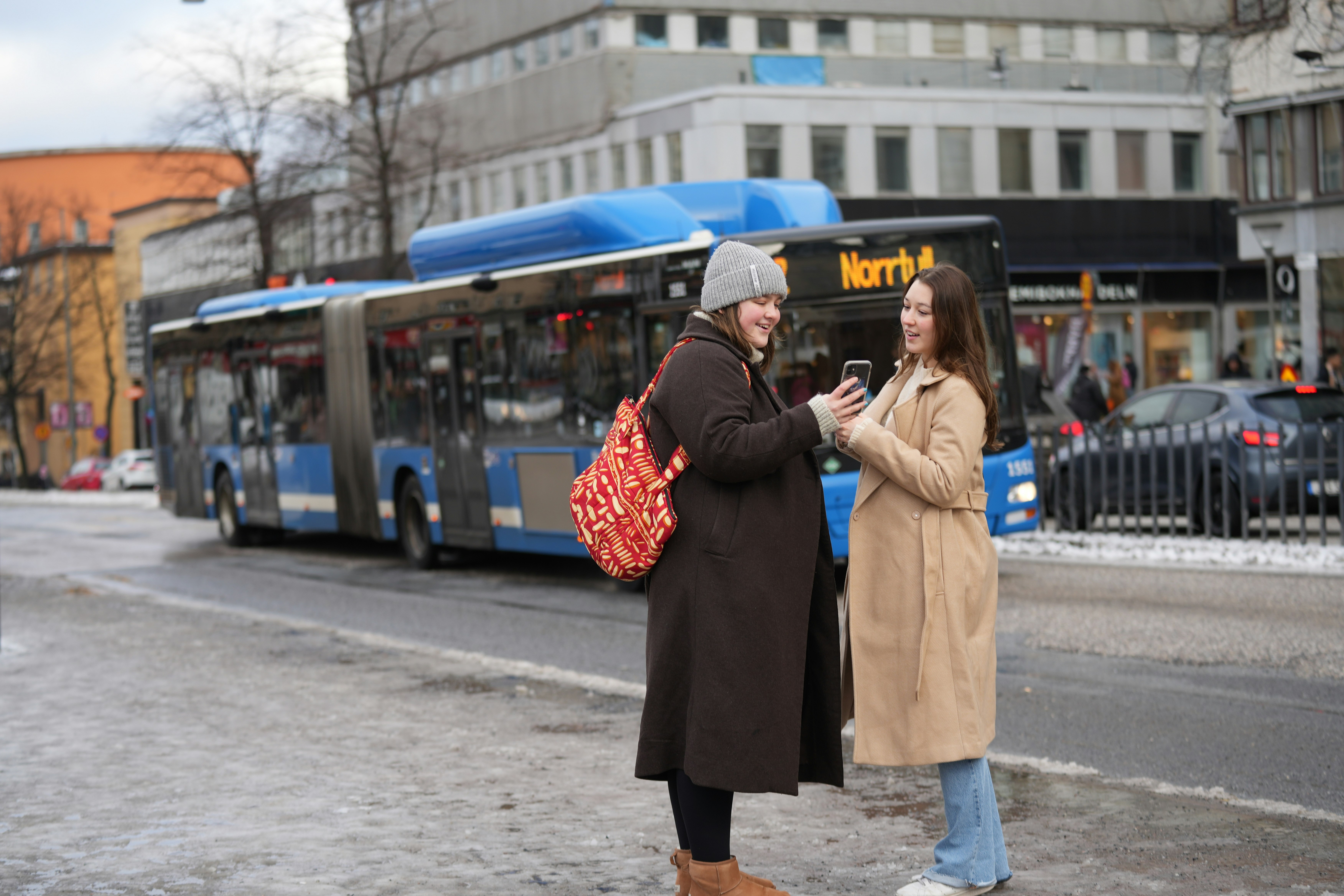 Two women chatting, their cell phone in hand, in front of a bus stop
