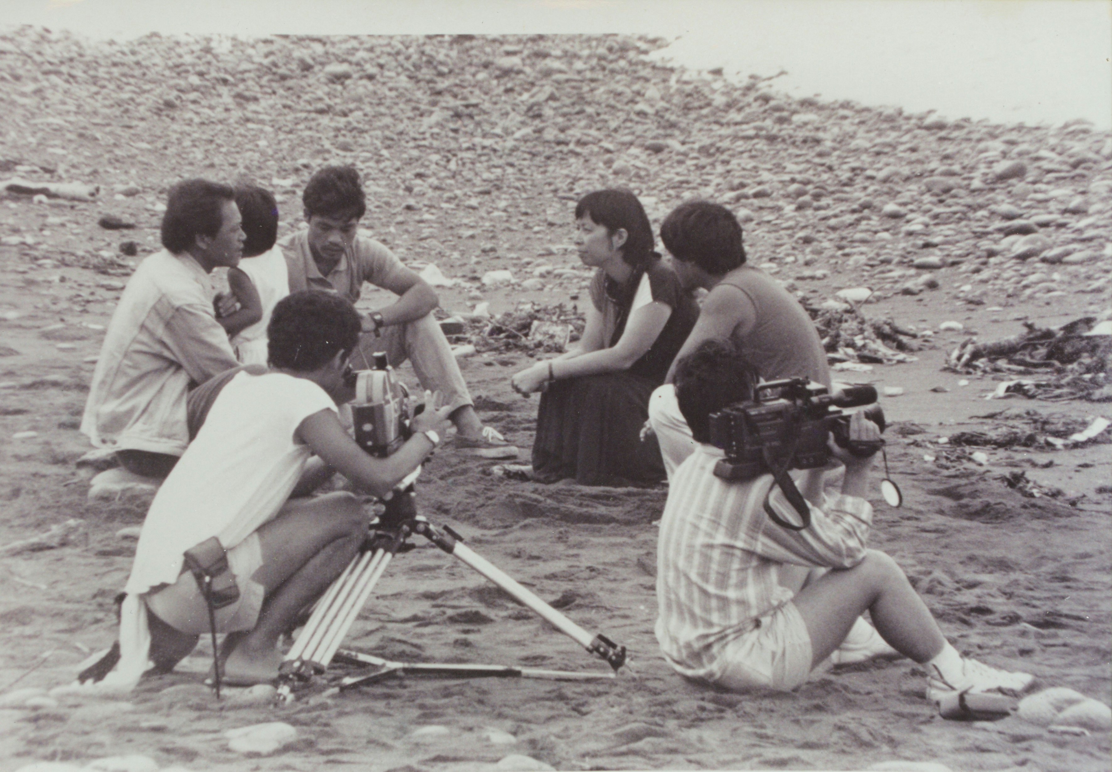 A group of people are sitting and kneeling down in a field. Two people are carrying camera equipment.