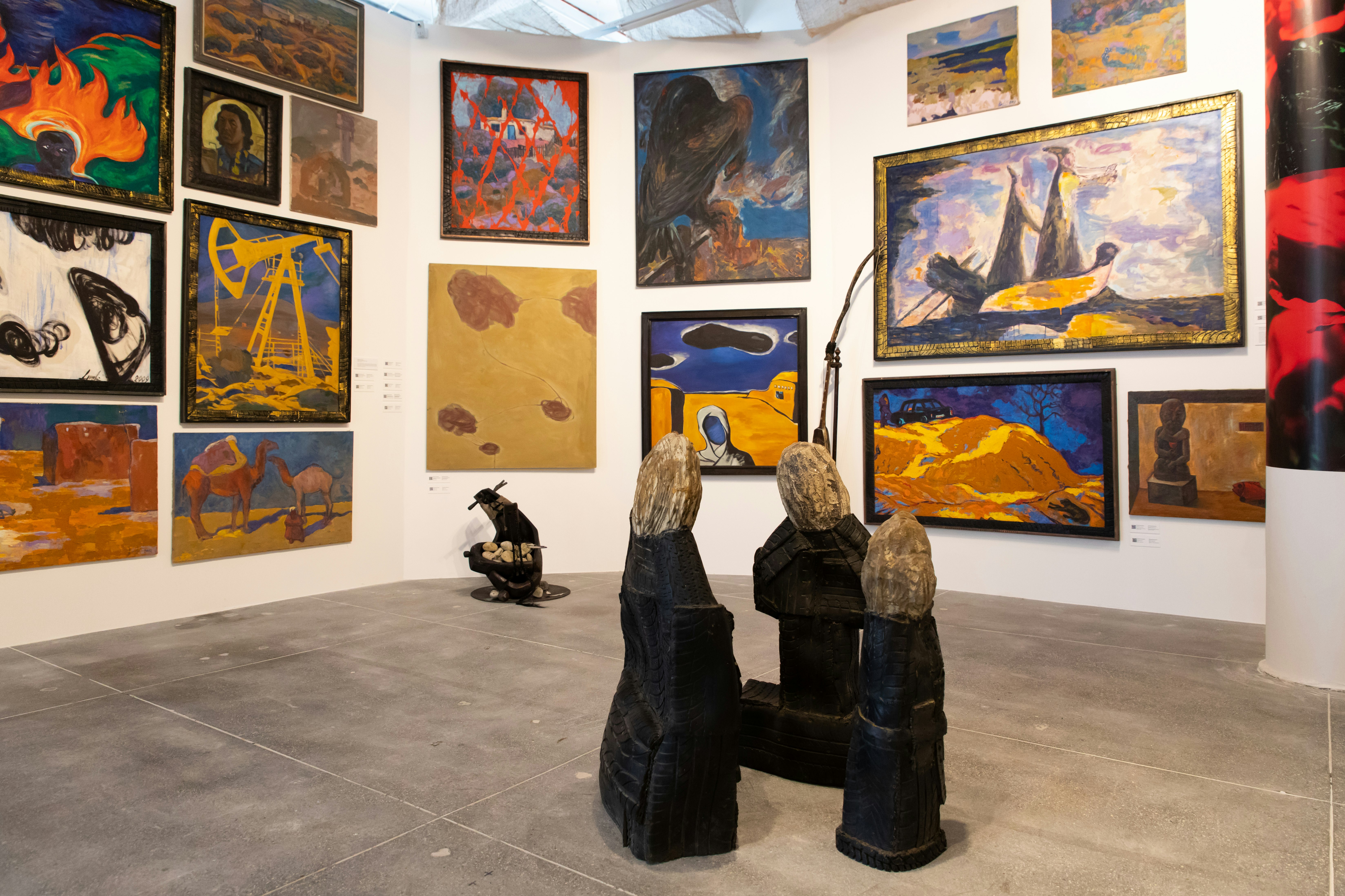 A sculpture resembling three figures facing eachother is in the foreground. In the background is a collection of artworks hung across three walls.