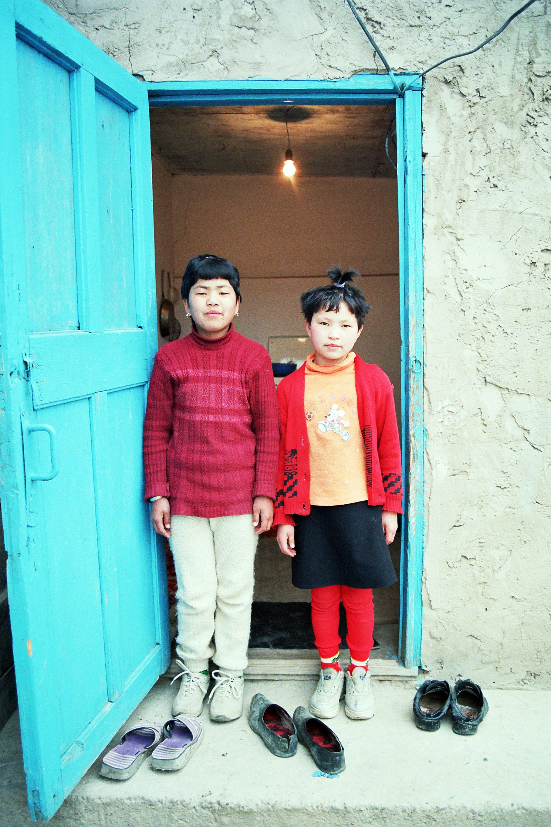 Two children standing in a blue doorway in front of three pairs of shoes.
