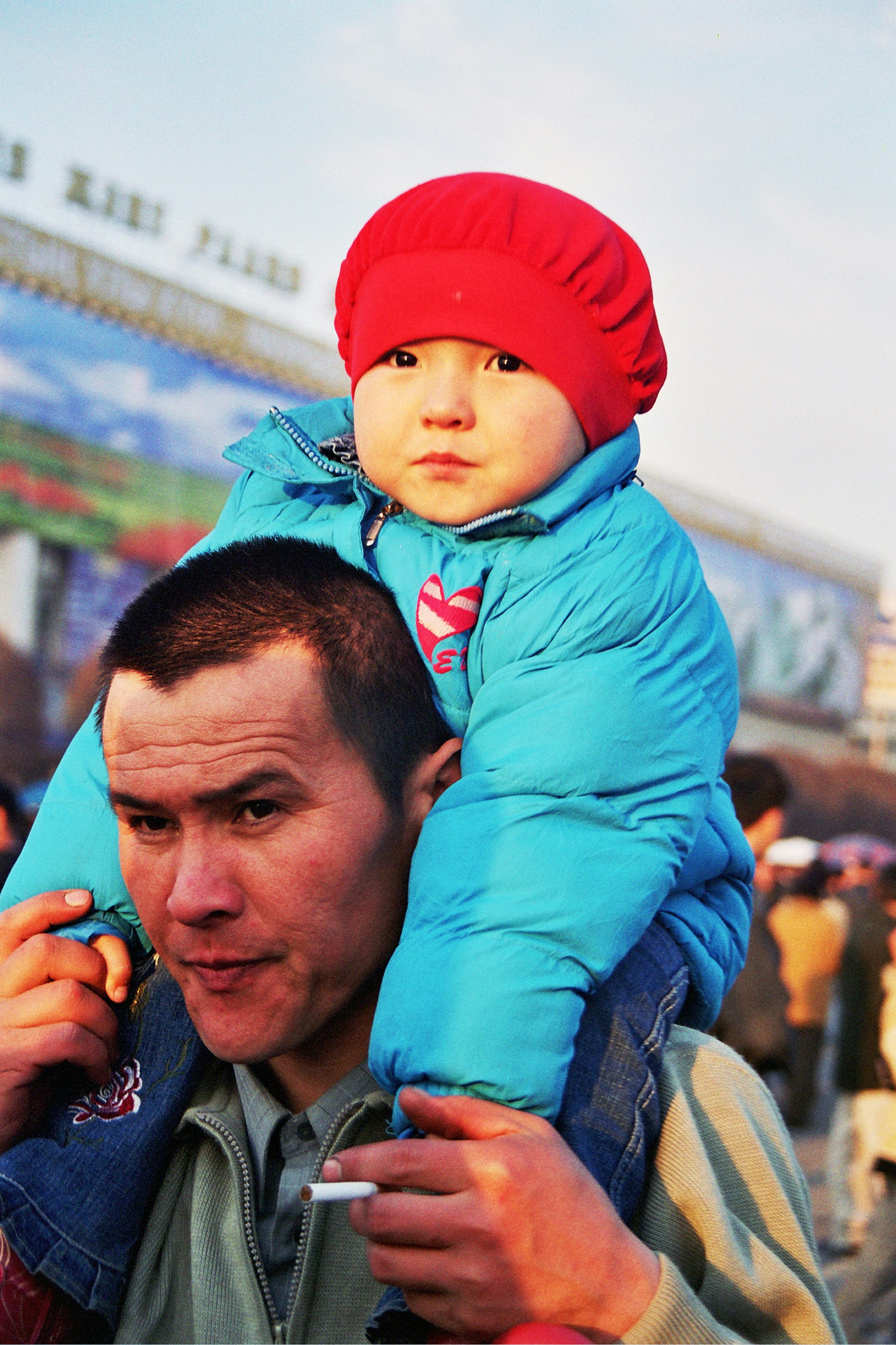 A small child in a blue jacket and a red hat on an adult's shoulders. The adult has a cigarette in their left hand and is holding the child's hand in their right.