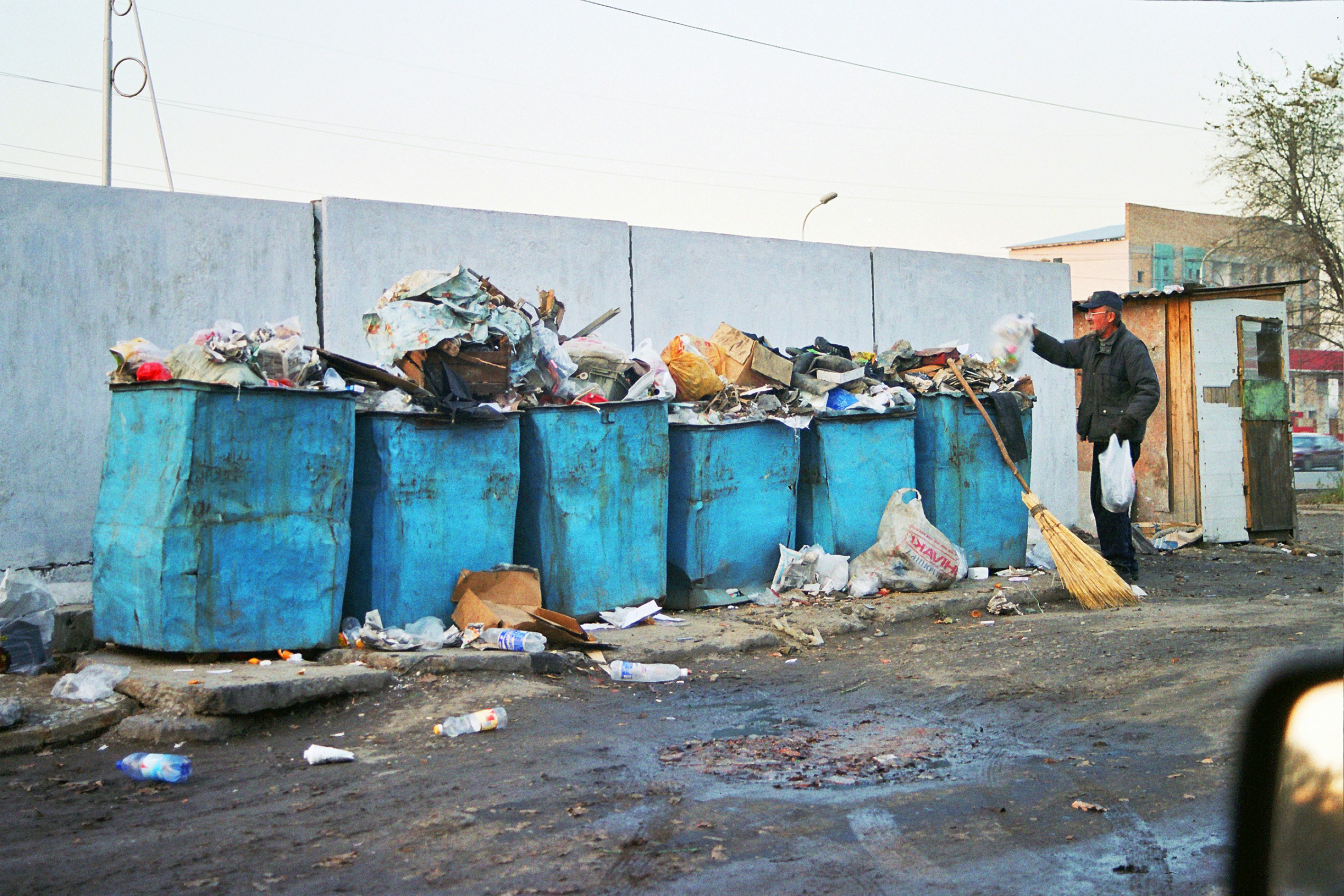 A line of six large blue bins filled to the brim with rubbish. An adult is pictured on the far right placing a bag in the bin.