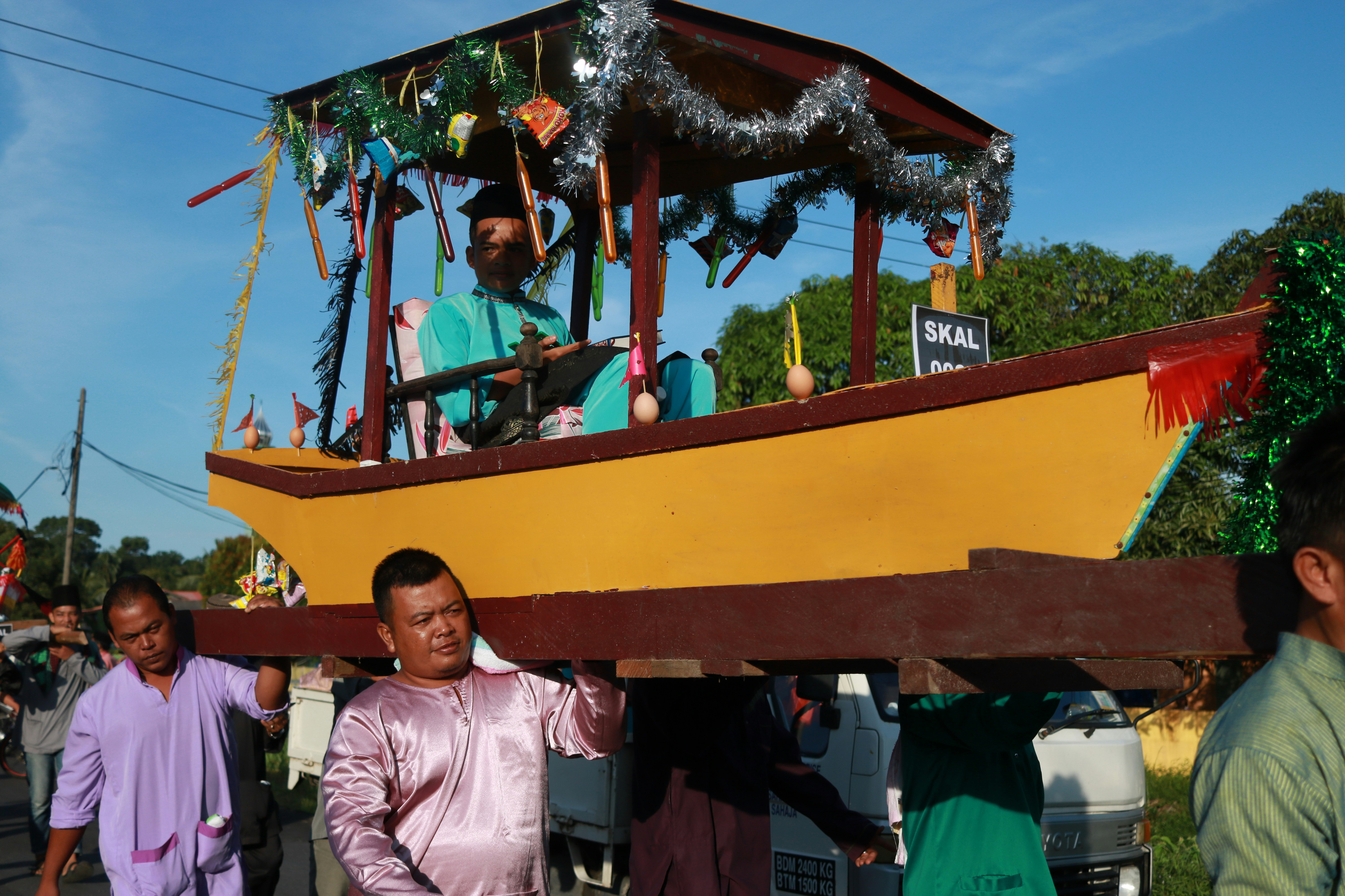 Men carrying a yellow boat decoated with silver tinsel. In the boat is a person seated and dressed in green.