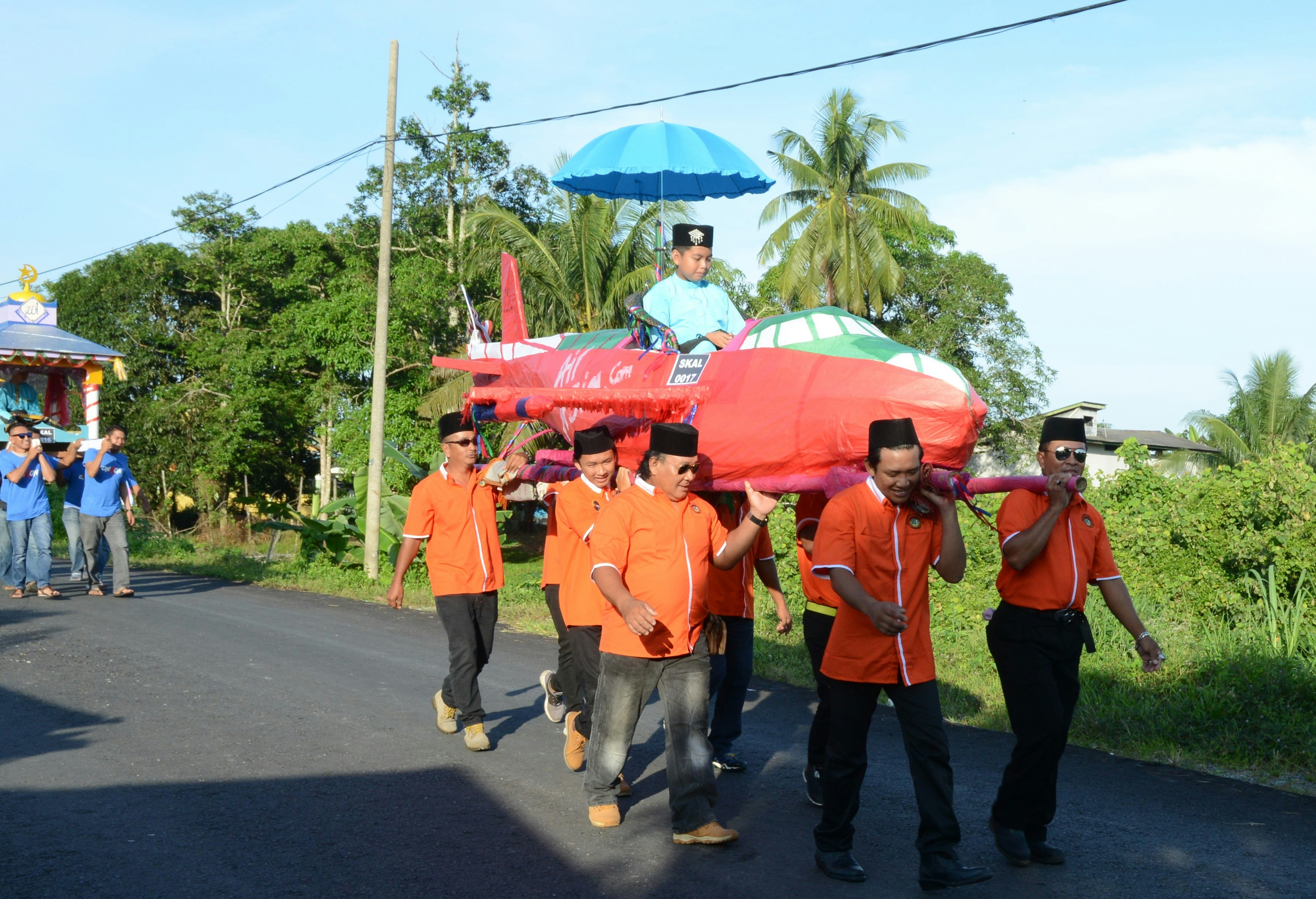 A group of men dressed in orange hold an usungan aircraft with a child dressed in blue in the passenger's seat.